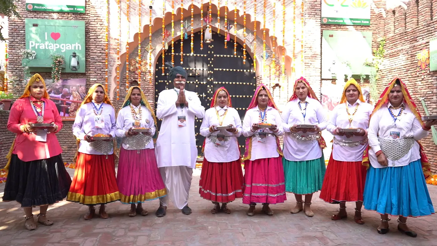 A man in white greeting with folded hands, flanked by women in traditional dress holding small bowls, at the decorated entrance to Pratapgarh Farms
